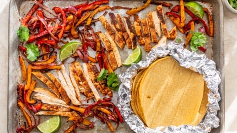 Sheet pan chicken fajitas with sliced cooked chicken, sautéed bell peppers, lime wedges, cilantro, and corn tortillas wrapped in foil.