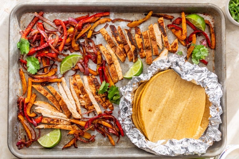 Sheet pan chicken fajitas with sliced cooked chicken, sautéed bell peppers, lime wedges, cilantro, and corn tortillas wrapped in foil.