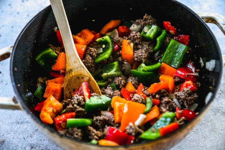 A pot with browned ground beef, chopped bell peppers, and onions being stirred with a wooden spoon.