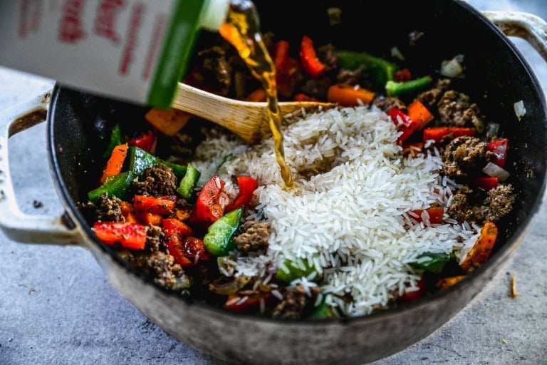 Broth is being poured into a pot with rice, ground meat, bell peppers, and onions, stirred with a wooden spoon.