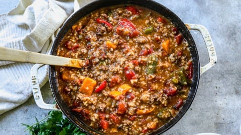 A pot of stuffed pepper soup with ground meat, beans, and colorful bell peppers, with a wooden spoon and herbs beside it.