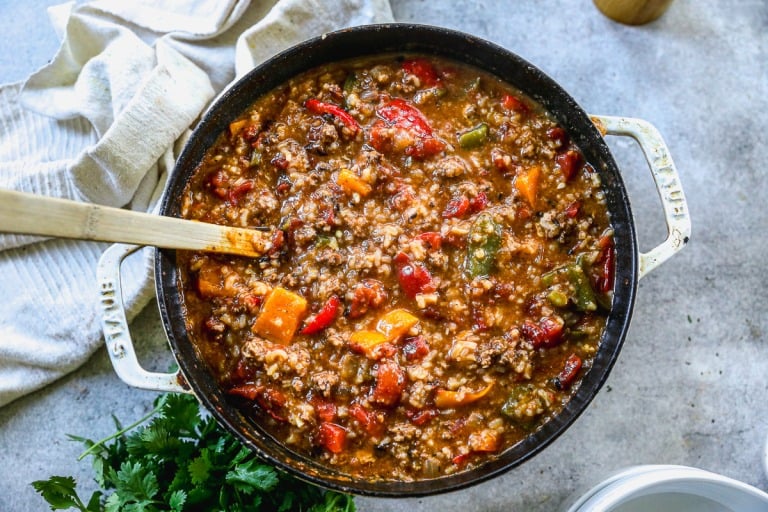A pot of stuffed pepper soup with ground meat, beans, and colorful bell peppers, with a wooden spoon and herbs beside it.