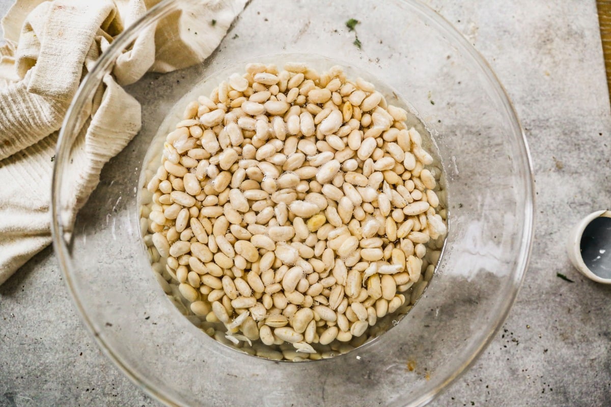 A bowl of white beans soaking in water on a gray countertop next to a cloth and a small empty cup.