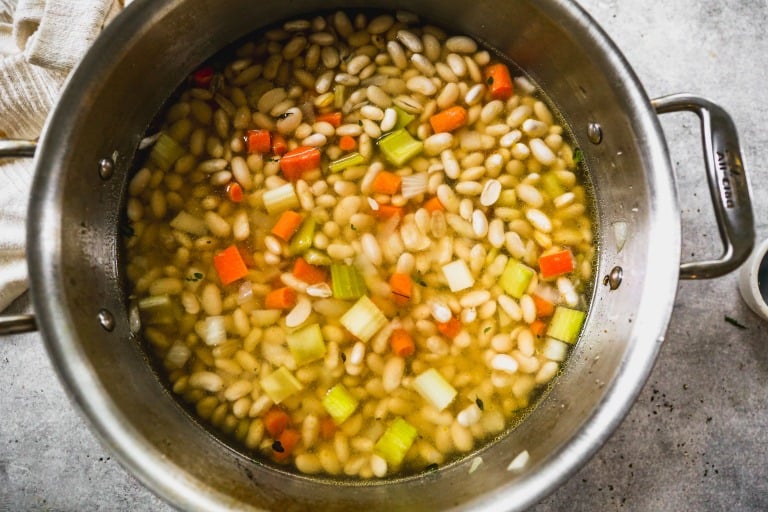 A pot of white bean soup with diced carrots and celery in a clear broth, viewed from above.