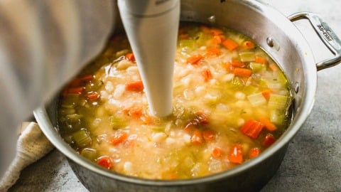 A hand uses an immersion blender to blend a pot of chunky white bean soup.