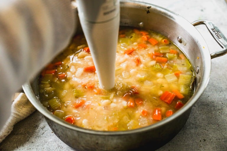 A hand uses an immersion blender to blend a pot of chunky white bean soup.