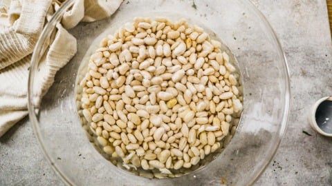 A bowl of white beans soaking in water on a gray countertop next to a cloth and a small empty cup.