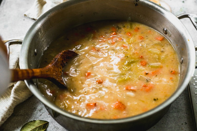 A pot of creamy white bean soup with carrots and celery being stirred with a wooden spoon.