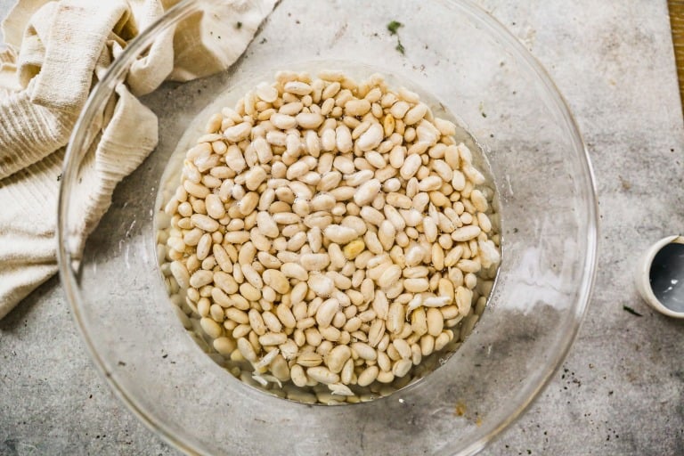 A bowl of white beans soaking in water on a gray countertop next to a cloth and a small empty cup.