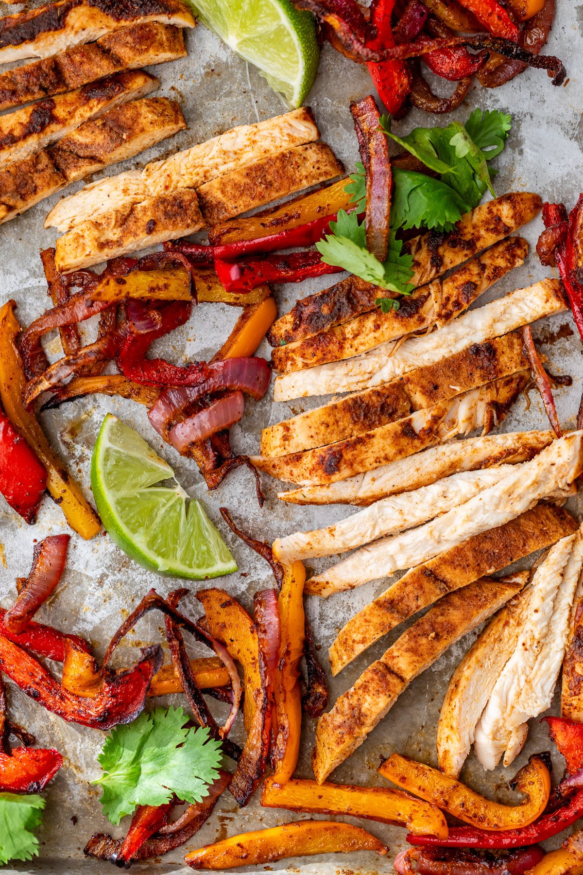 Sheet pan chicken fajitas with red and yellow bell peppers, onions, lime wedges, and cilantro on a tray.