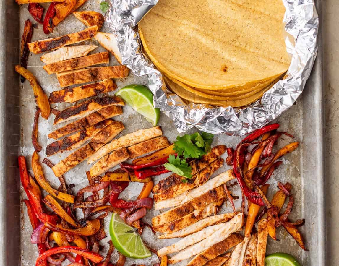 Sheet pan chicken fajitas with sliced seasoned chicken, sautéed peppers and onions, lime wedges, and corn tortillas wrapped in foil.