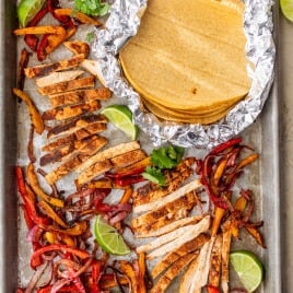Sheet pan chicken fajitas with sliced seasoned chicken, sautéed peppers and onions, lime wedges, and corn tortillas wrapped in foil.