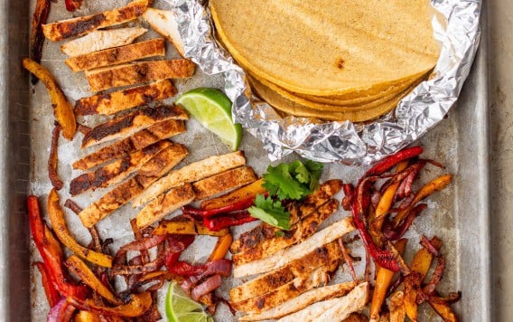 Sheet pan chicken fajitas with sliced seasoned chicken, sautéed peppers and onions, lime wedges, and corn tortillas wrapped in foil.