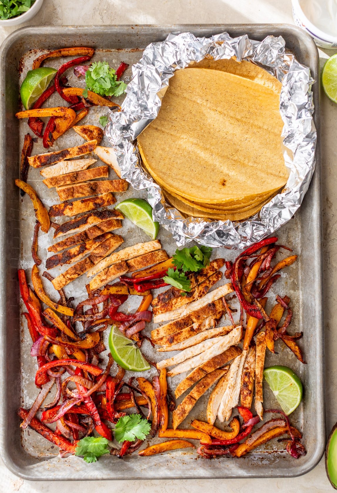 Sheet pan chicken fajitas with sliced seasoned chicken, sautéed peppers and onions, lime wedges, and corn tortillas wrapped in foil.
