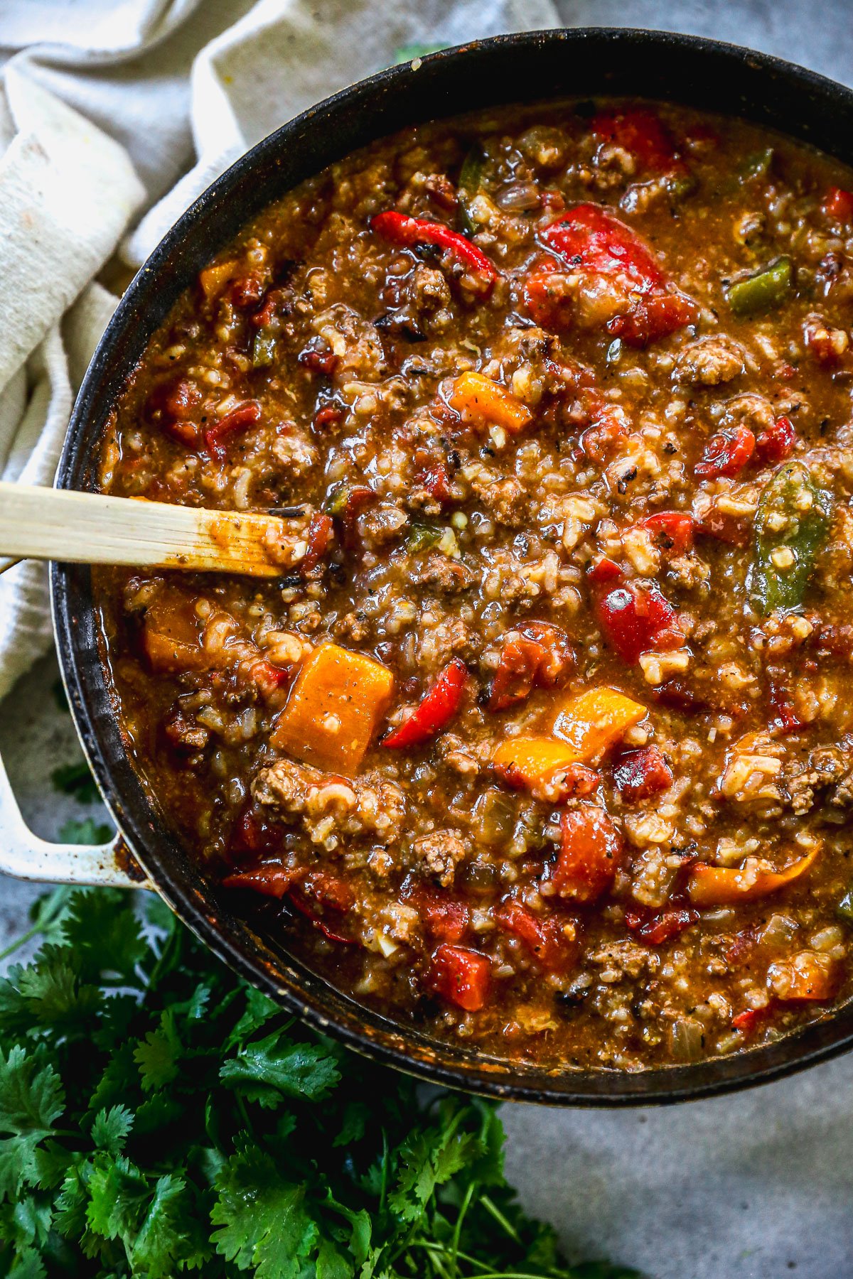 A pot of hearty stuffed pepper soup with ground meat, bell peppers, tomatoes, and rice, next to fresh cilantro and a wooden spoon.
