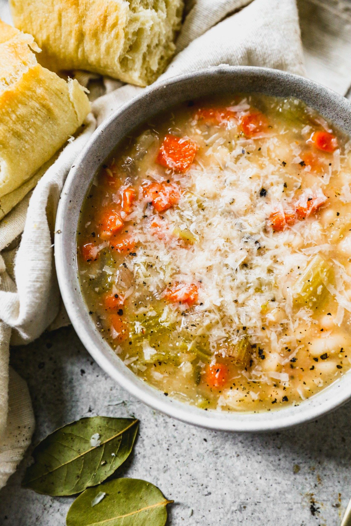 A bowl of white bean soup topped with grated cheese, next to torn bread and bay leaves on a gray surface.