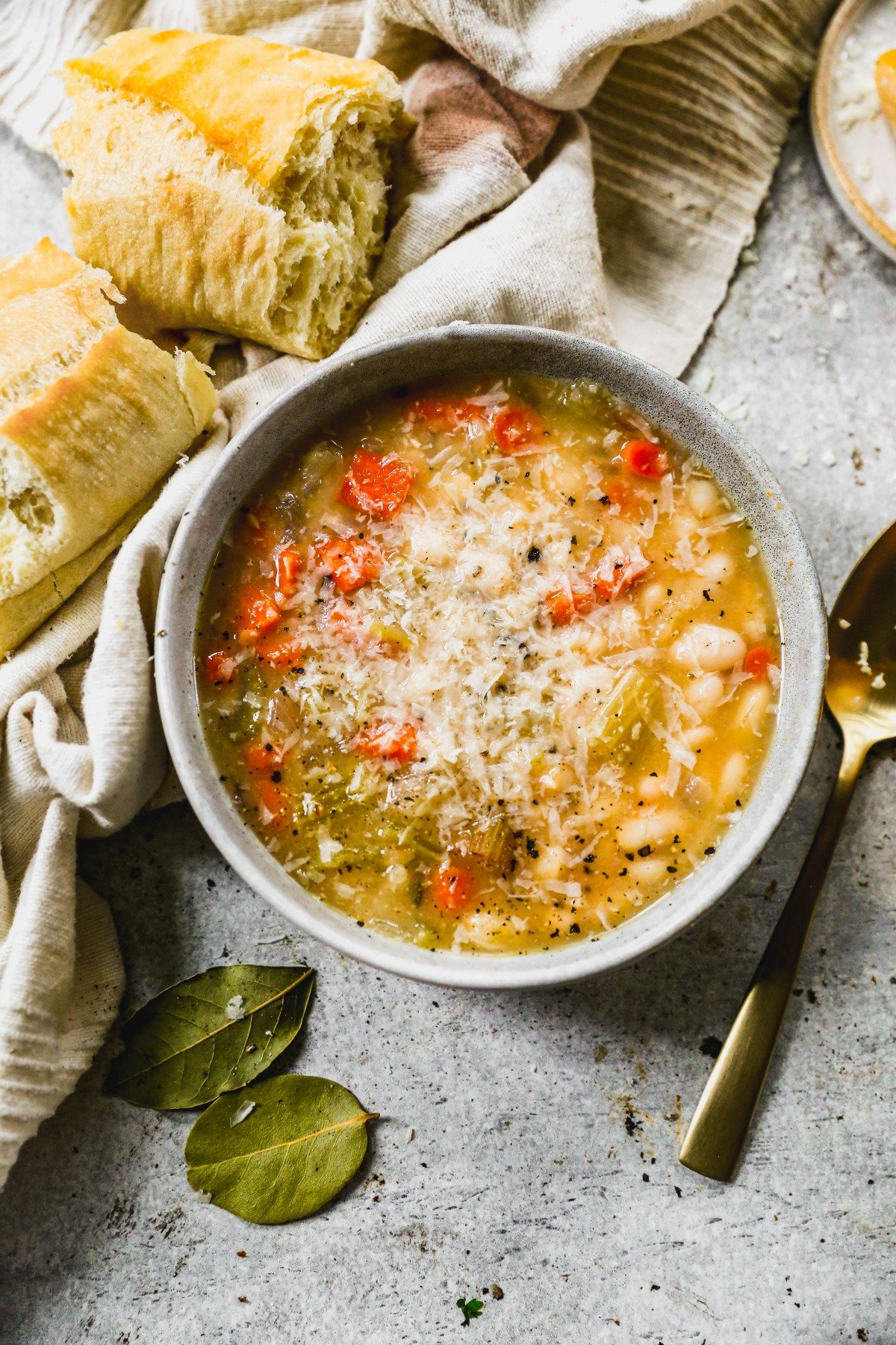 A bowl of white bean soup with grated cheese, served with crusty bread, a spoon, and bay leaves on the side.