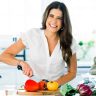 Smiling woman in a white shirt chops a red bell pepper on a cutting board with vegetables in a bright kitchen.