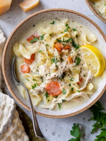 A bowl of creamy turkey noodle soup with carrots, parsley, a lemon wedge, and a spoon on the side.