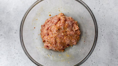 Ground pork mixed with spices in a clear glass bowl on a light gray surface.