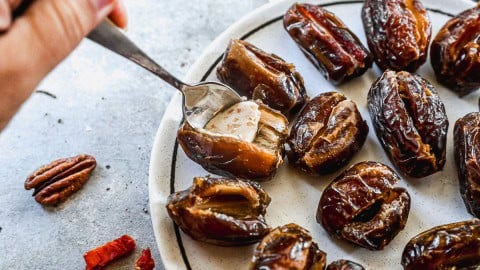 A hand stuffs a date with a creamy filling on a plate of dates, pecans, and spices nearby.