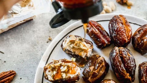 A plate of dates stuffed with cream, seeds, nuts, and drizzled with honey, surrounded by nuts on a gray surface.