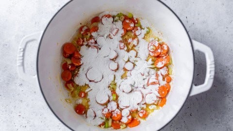 Sliced carrots, celery, and onions in a white pot, topped with a layer of flour, on a light surface.