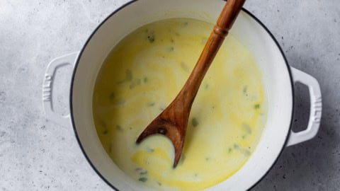 A pot of creamy yellow soup with green herbs, stirred by a wooden spoon on a light gray surface.