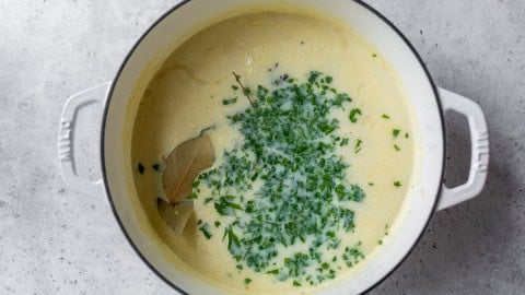 Creamy soup in a white pot, topped with chopped herbs, bay leaves, and a splash of milk on a gray surface.
