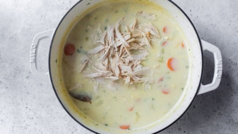 A pot of creamy turkey soup with shredded turkey, carrots, and herbs on a light gray surface.