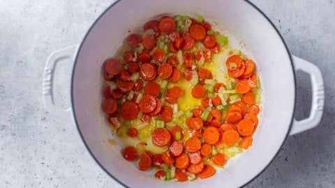 Sliced carrots, celery, and onions sautรฉing in a white pot on a gray countertop.