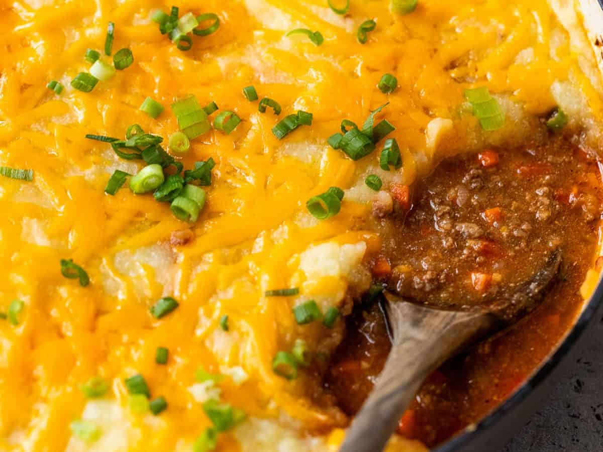A skillet of sloppy joe casserole topped with melted cheddar and chopped green onions, with a wooden spoon scooping some out.