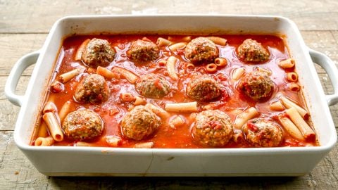 White baking dish with meatballs, pasta, and tomato sauce on a wooden surface.