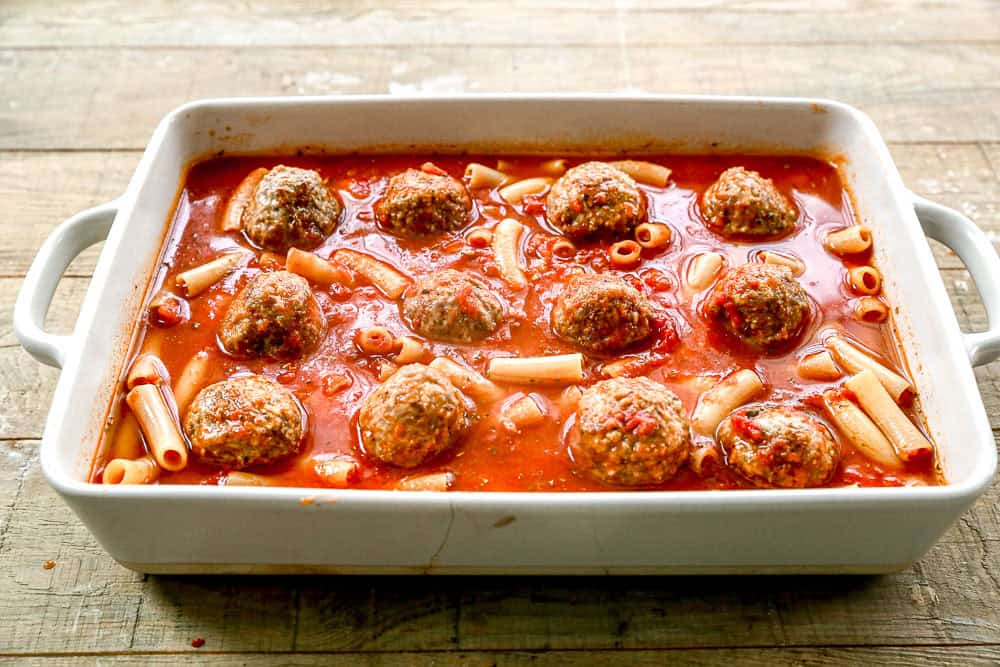 White baking dish with meatballs, pasta, and tomato sauce on a wooden surface.
