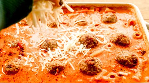 A hand sprinkles shredded cheese over meatballs in a red sauce in a white baking dish.