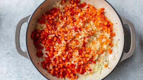 Chopped onions, red bell peppers, and carrots sautéing in a large pot on a light surface.