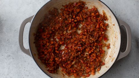 Chopped onions, carrots, and red peppers sautéing in a round white pot on a light gray surface.