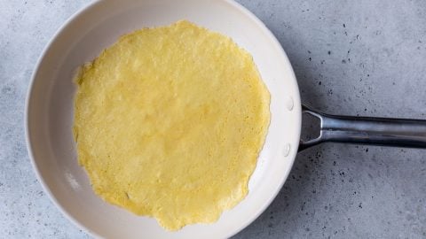 A thin yellow Swedish pancake cooking in a white frying pan on a light gray countertop.