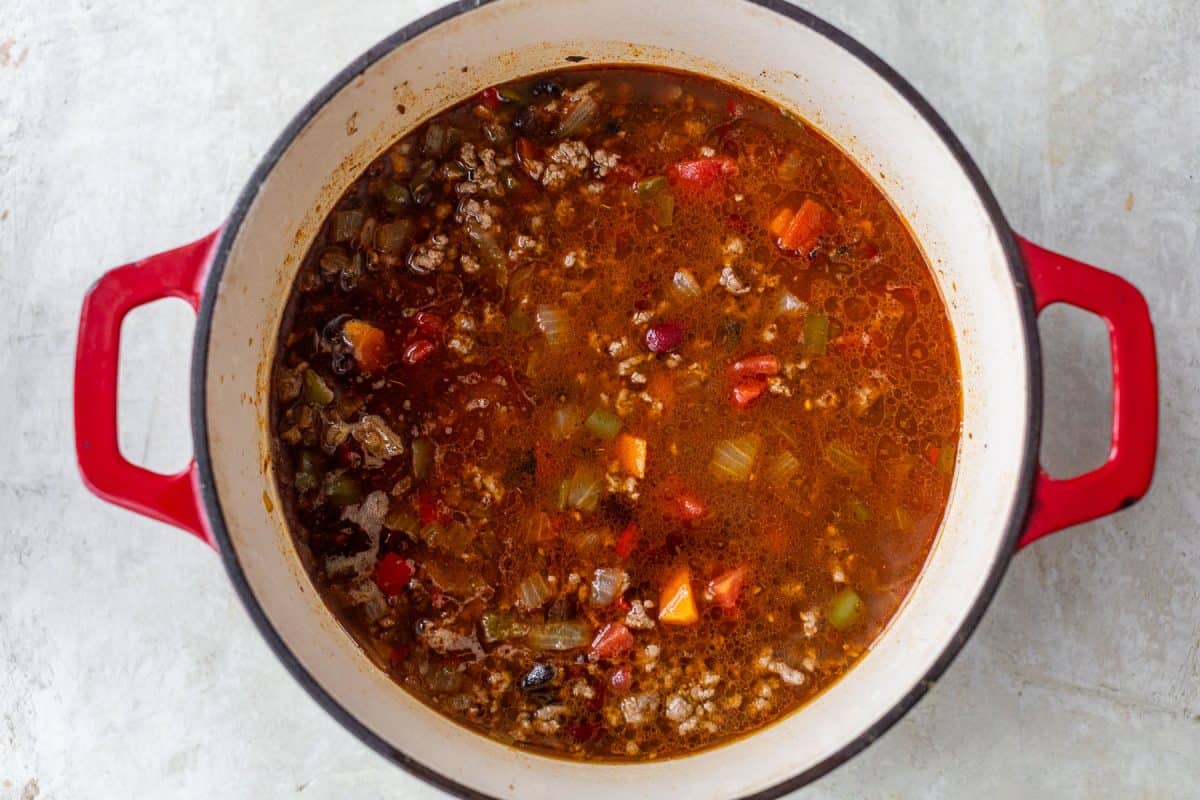 A pot of hearty soup with ground meat, beans, and vegetables, in a red Dutch oven on a light surface.