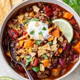 A bowl of ground beef chili with beans, vegetables, cheese, sour cream, tortilla chips, and a lime wedge, with a spoon inside.