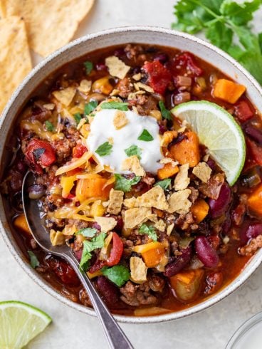 A bowl of ground beef chili with beans, vegetables, cheese, sour cream, tortilla chips, and a lime wedge, with a spoon inside.