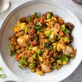 A bowl of quinoa fried rice with green onions, scrambled eggs, and vegetables on a light-colored surface.