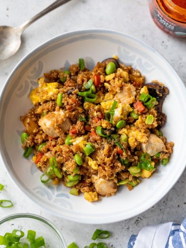 A bowl of quinoa fried rice with green onions, scrambled eggs, and vegetables on a light-colored surface.