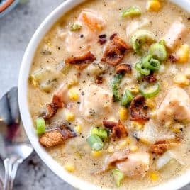 A bowl of creamy salmon chowder soup with chunks of salmon, bacon, corn, potatoes, and green onions, with spoons beside it.