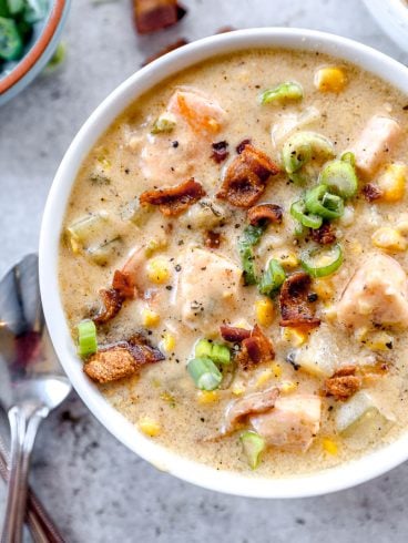 A bowl of creamy salmon chowder soup with chunks of salmon, bacon, corn, potatoes, and green onions, with spoons beside it.