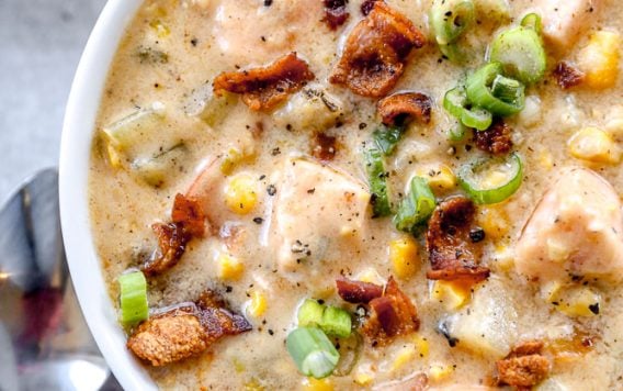 A bowl of creamy salmon chowder soup with chunks of salmon, bacon, corn, potatoes, and green onions, with spoons beside it.