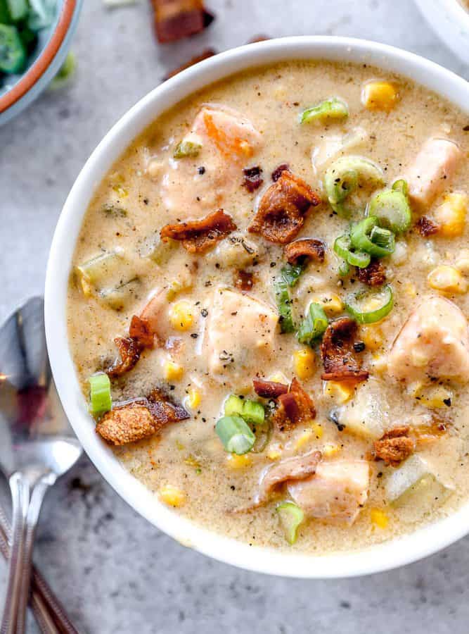 A bowl of creamy salmon chowder soup with chunks of salmon, bacon, corn, potatoes, and green onions, with spoons beside it.