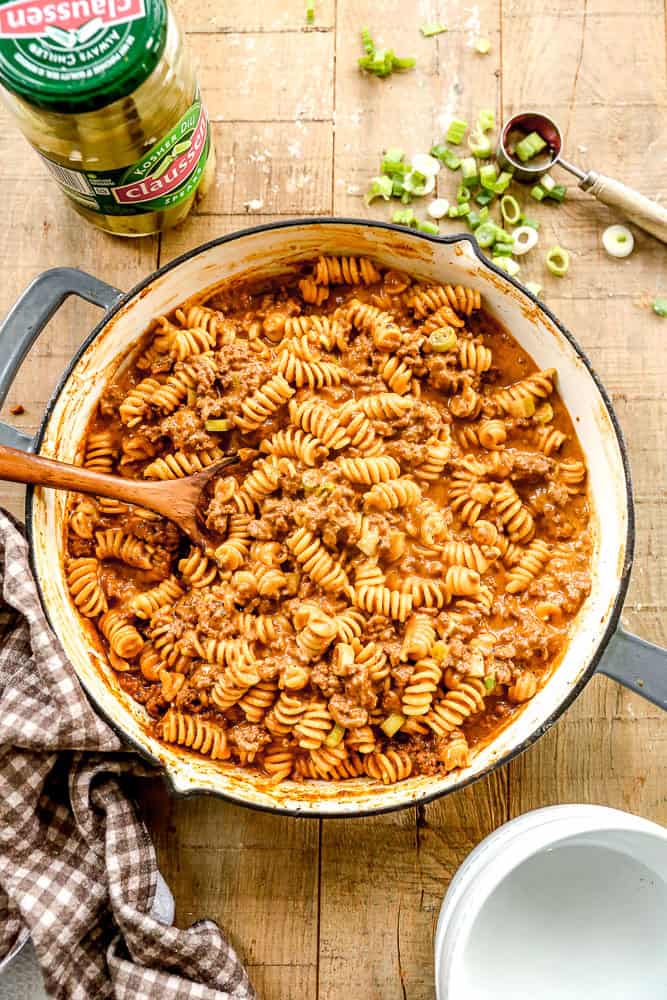 A pot of cheeseburger casserole with ground beef and sauce, topped with sliced green onions, sits on a wooden table.