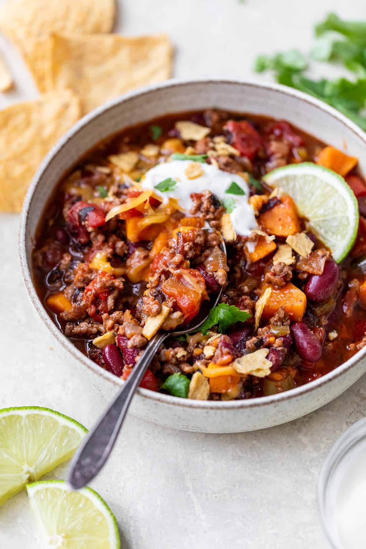 A bowl of ground beef chili topped with sour cream, lime, and herbs, with a spoon inside. Tortilla chips and lime slices nearby.