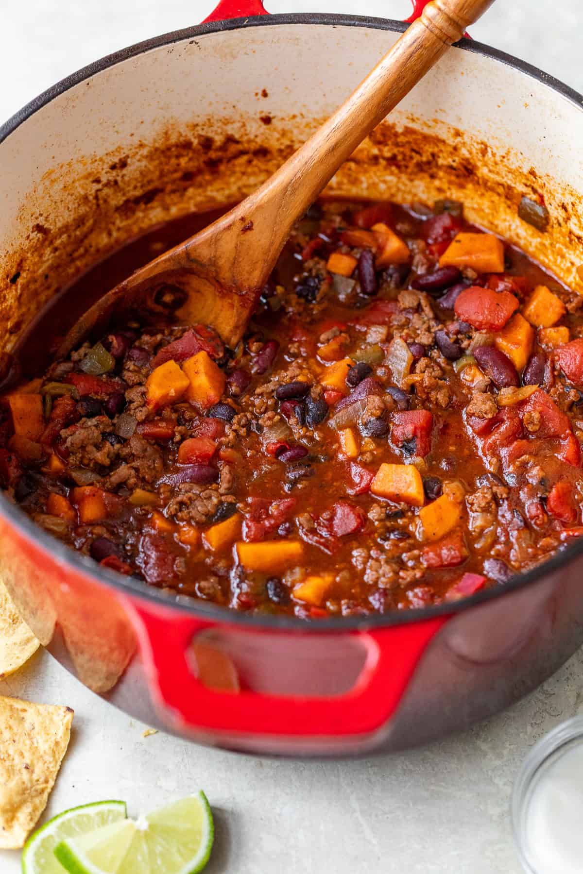 A red pot filled with ground beef chili containing beans, tomatoes, and sweet potatoes, with a wooden spoon inside.